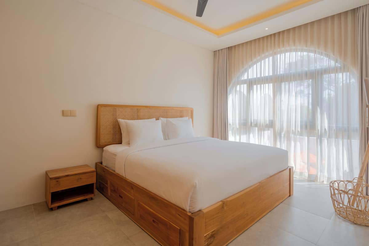 A bedroom with a wooden bed, a nightstand, a wicker chair, and a large window with sheer curtains.