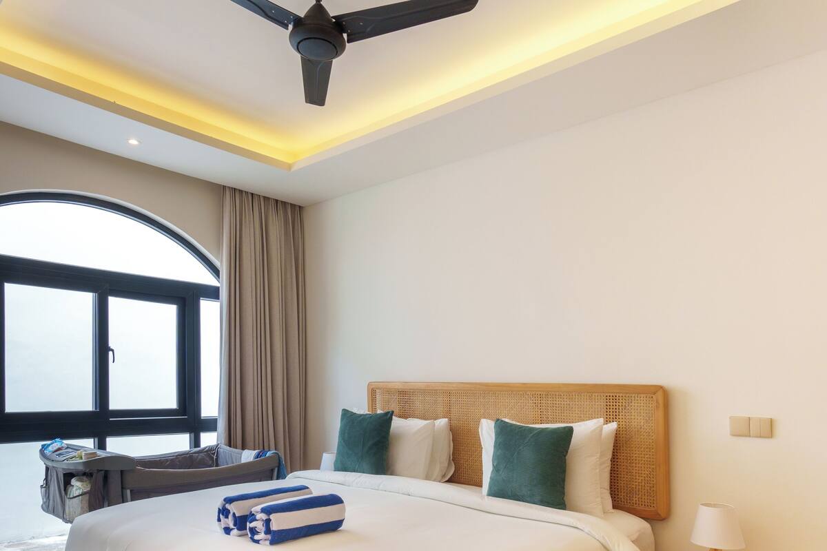 A modern hotel room with a large bed, a ceiling fan, a wooden headboard, and a view of the outdoors.