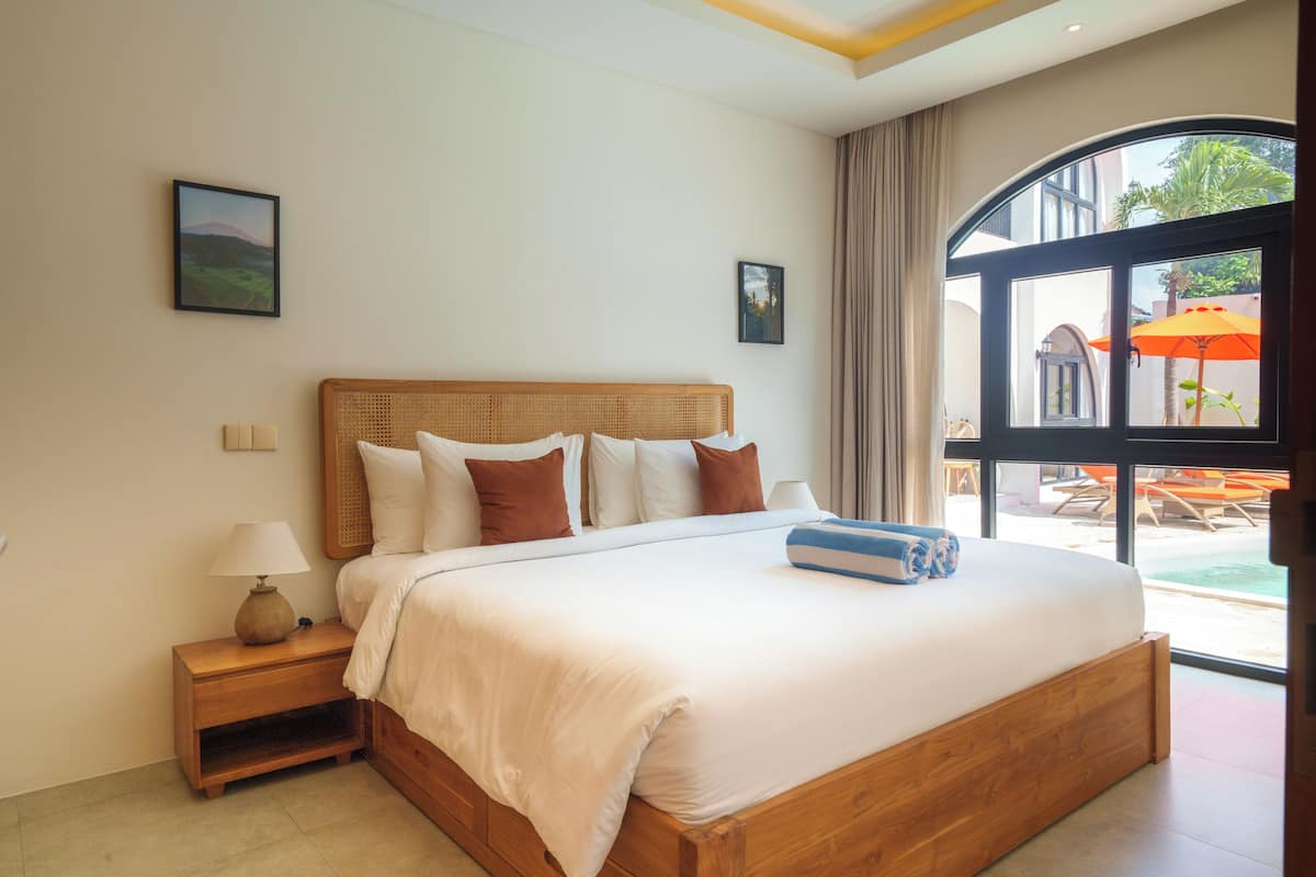A large bed with a wooden headboard, white bedding, and multiple pillows. There is a nightstand with a lamp and a small shelf. A window with curtains and an outdoor view is visible.