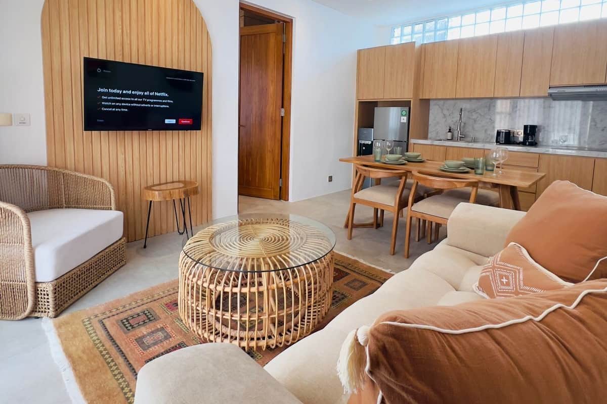 A modern living room with a flat-screen TV, a ceiling fan, and a kitchen area with wooden cabinets and a dining table.