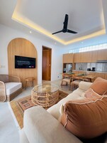 A modern living room with a flat-screen TV, a ceiling fan, and a dining area with a kitchen in the background.