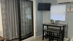 Un espace de vie compact doté d’une porte-fenêtre coulissante, d’un téléviseur à écran plat fixé au mur, d’une petite table à manger avec deux chaises et d’un canapé agrémenté d’un plaid à motifs.
