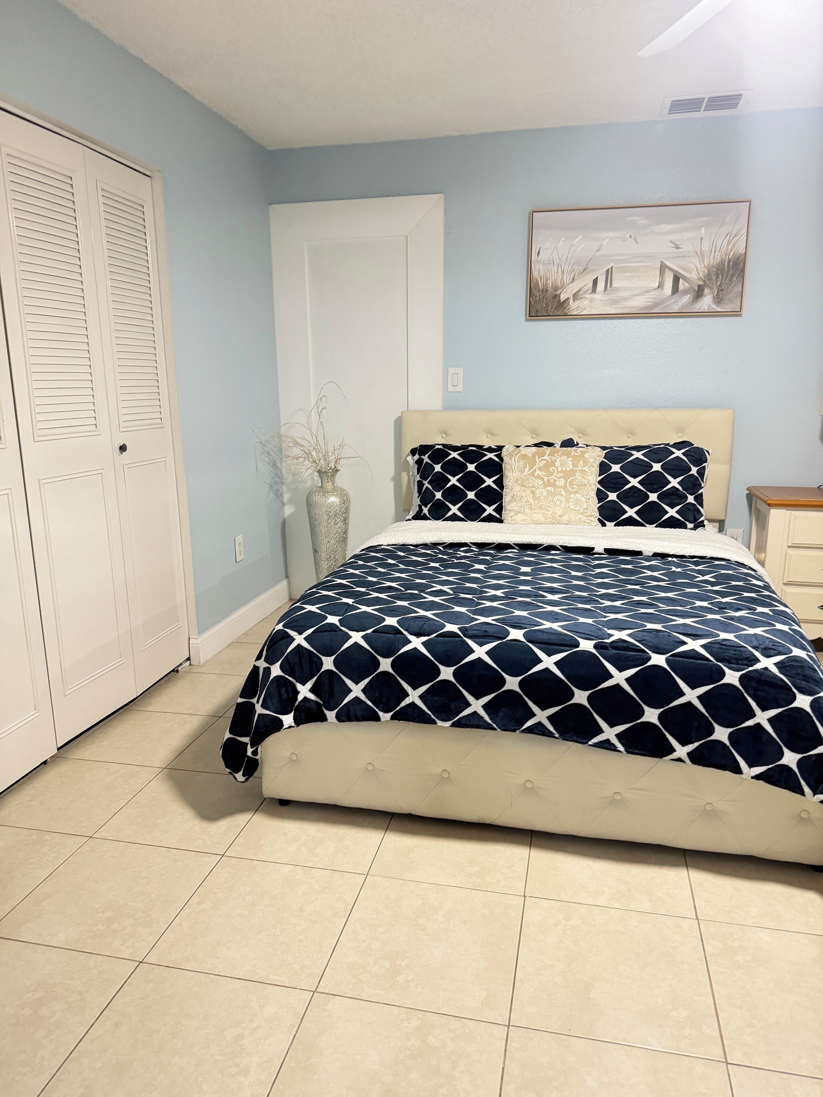 A bedroom with a bed, a nightstand, a vase with plants, a door, and a painting on the wall.