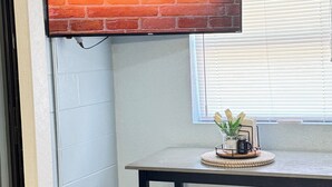 Une pièce avec un panneau néon indiquant « Bienvenue » sur un mur de briques, une fenêtre avec des stores, une table sur laquelle se trouve un plateau rempli d’objets et un banc.