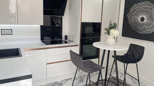 A modern kitchen with white cabinetry, black appliances, and a marble floor. There is a dining area with two chairs and a small table. - Modern-luxury 1bedroom-EnsuiteBathroom-Garden-Wifi (Basildon)