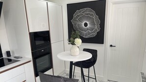 A modern kitchen with white cabinetry, a black oven, and a white dining table with two black chairs. A vase with flowers is on the table, and a black and white abstract painting is on the wall. - Modern-luxury 1bedroom-EnsuiteBathroom-Garden-Wifi (Basildon)
