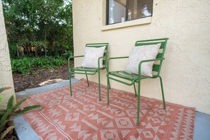 Deux chaises en métal vert, dotées de coussins à motifs, sont posées sur un tapis à motifs, sur un sol en béton, contre un mur percé d’une fenêtre.