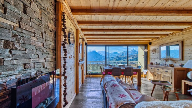 Een gezellig interieur van een chalet met een stenen muur, een open haard, een eethoek en uitzicht op bergen.
