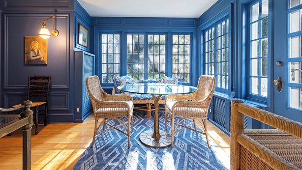 Una habitación con una mesa redonda de vidrio rodeada de sillas de mimbre, ventanas amplias con detalles azules y una alfombra estampada sobre el suelo de madera.