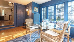 Una habitación con una mesa de vidrio rodeada de sillas de mimbre, una puerta azul y ventanas amplias.
