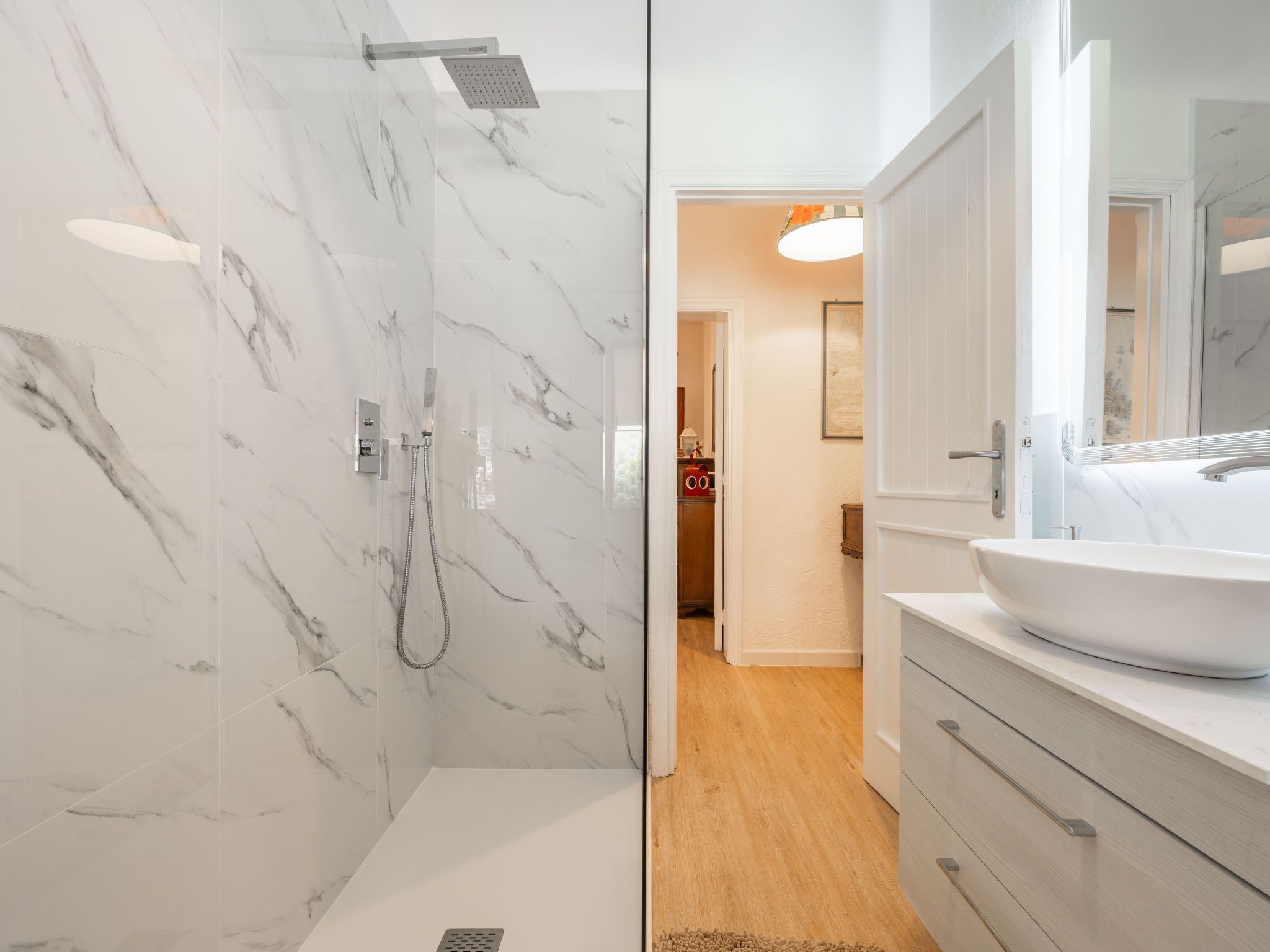 Une salle de bain moderne avec un mur de douche en marbre, un lavabo blanc et un meuble-lavabo en bois.