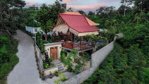 Ein Bungalow mit rotem Ziegeldach, einer Veranda mit Reetdach und einer Steinsäule, die das Grundstück umgibt.