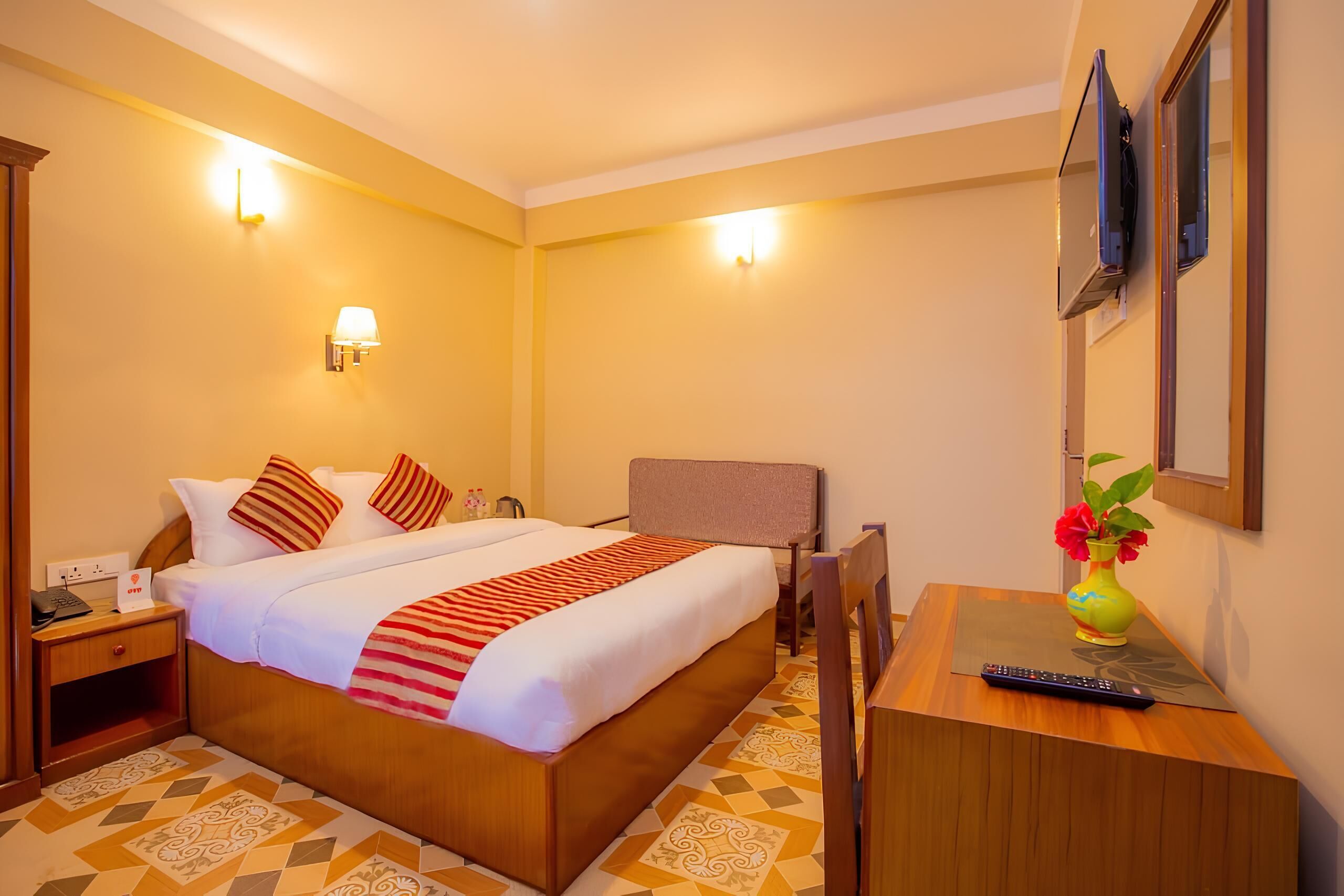Una habitación de hotel con cama, mesita de noche, lámpara y un jarrón con flores.