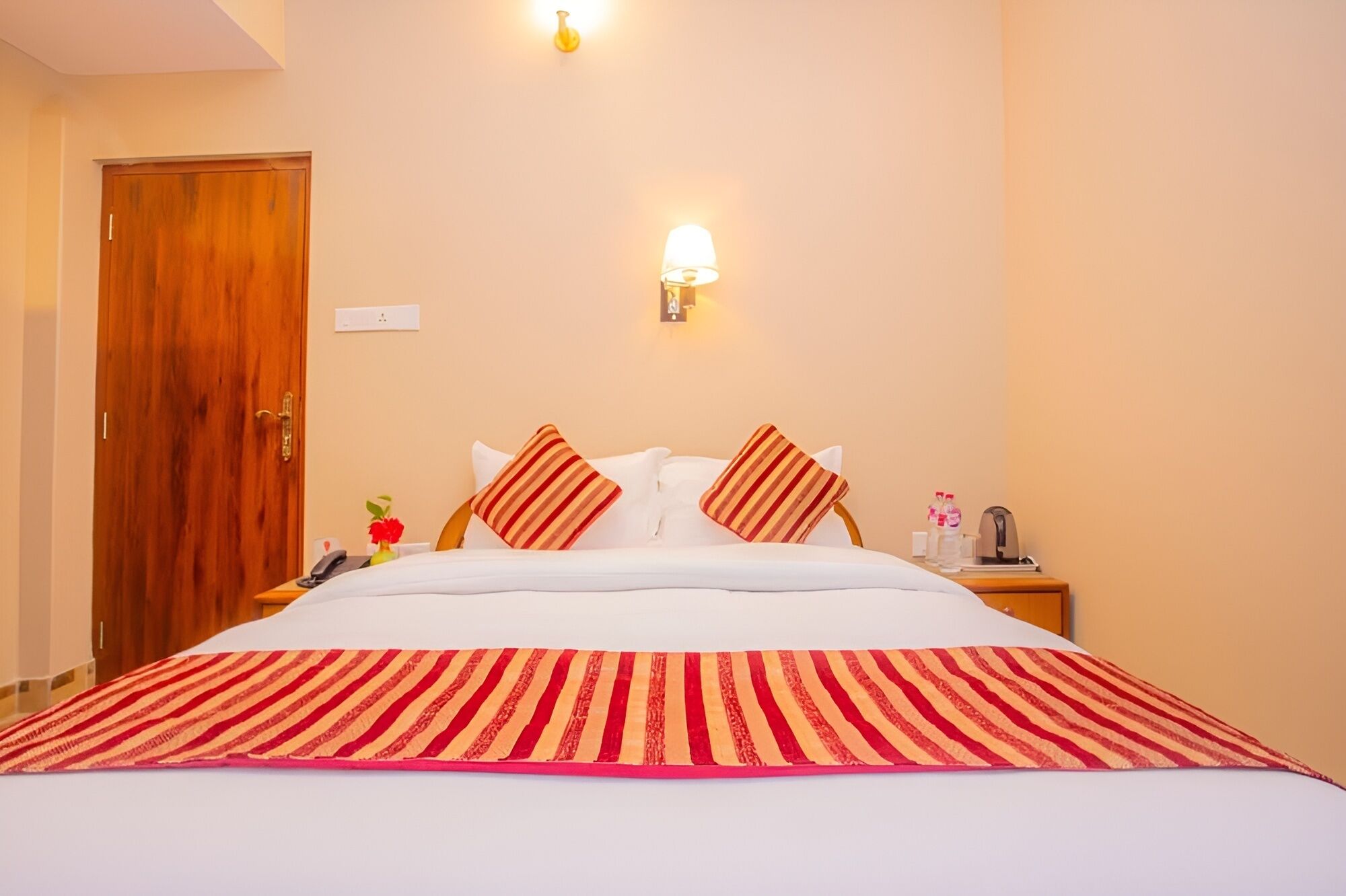 A hotel room with a large bed, a wooden door, a bedside table with a lamp, and a wall-mounted light fixture.