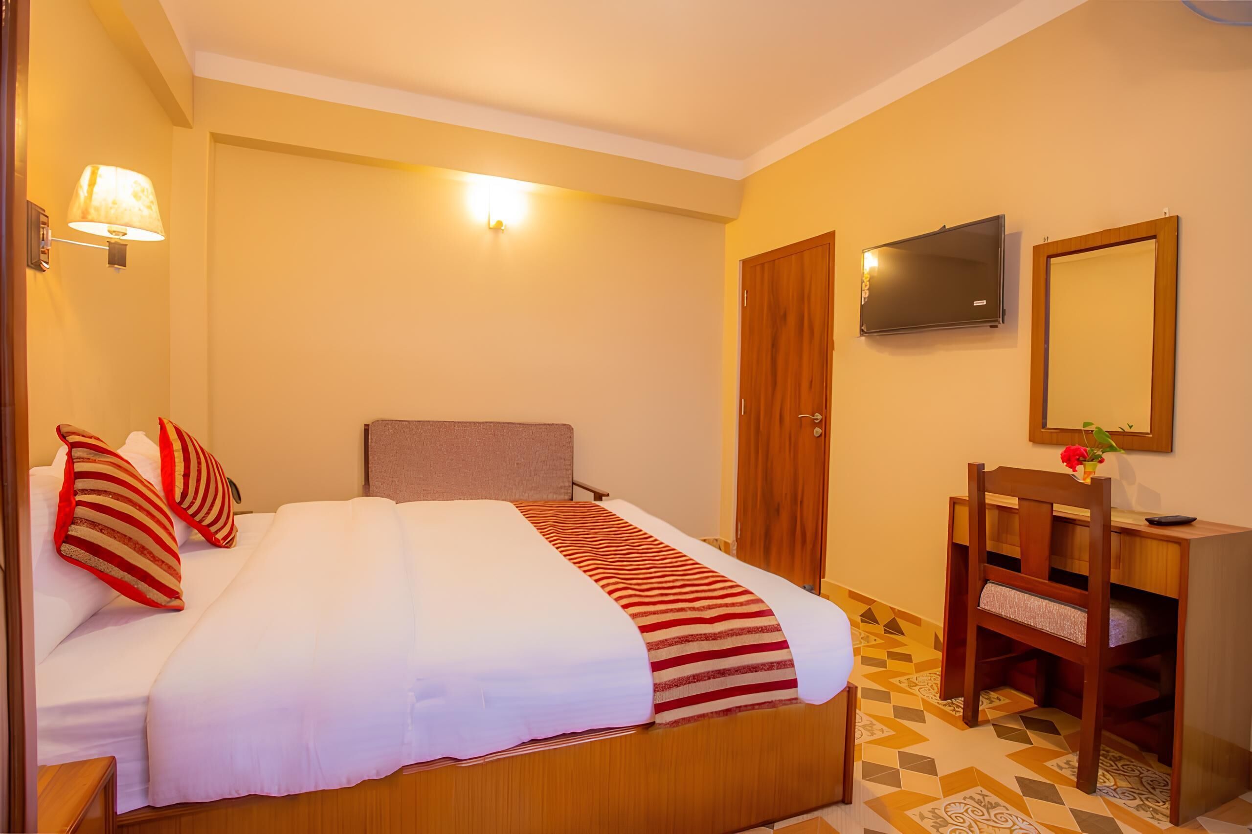 A hotel room with a bed, bedside lamp, TV, wooden desk, chair, and a mirror.