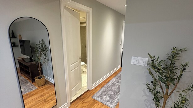 A hallway with a wooden floor, a mirror, a potted plant, and a door leading to a bathroom. - Beautiful Priv. bsmt 2B 1Bath apartment w/ parking (Brampton)
