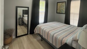 Un dormitorio con cama, mesita de noche, espejo, ventilador de techo y ventana con cortinas.