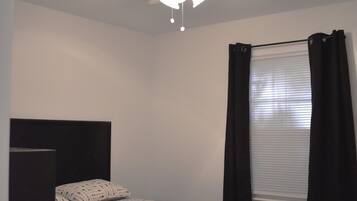 Un dormitorio con cama, ventilador de techo, ventana con cortinas y mesita de noche.