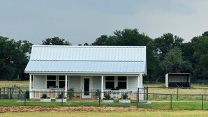 A single-story house with a gabled roof, a front porch, and a fenced yard. - Private, serene cottage on a horse ranch w/ spa vibes, near Granbury & Ft Worth. (Granbury)