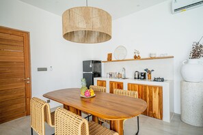 A dining area with a wooden table and chairs, a hanging light fixture, and a kitchen in the background. - Villa Madeleine Uluwatu (Pecatu)