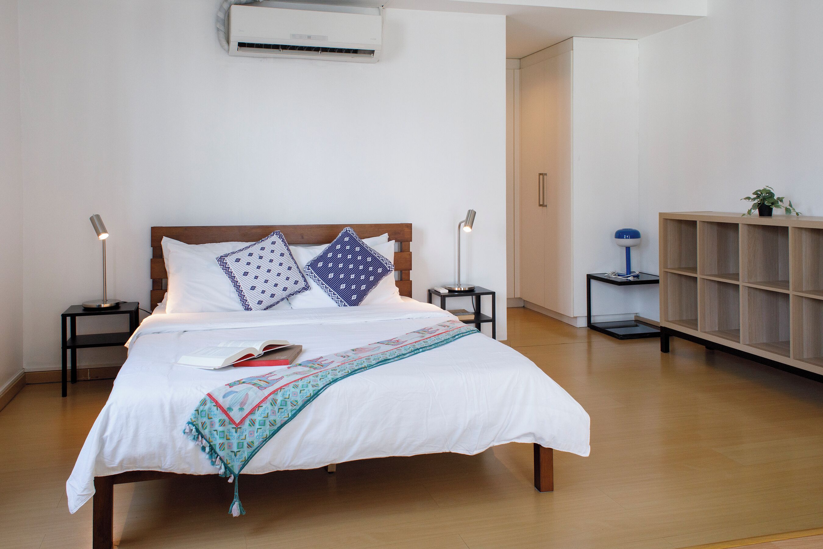 Una cama bien hecha con edredón blanco y almohadones decorativos, una mesita de noche con lámpara y una mesita auxiliar de madera.