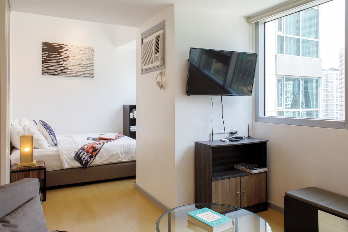 A modern hotel room with a bed, a glass coffee table with a book, a TV mounted on the wall, and a window with a city view.