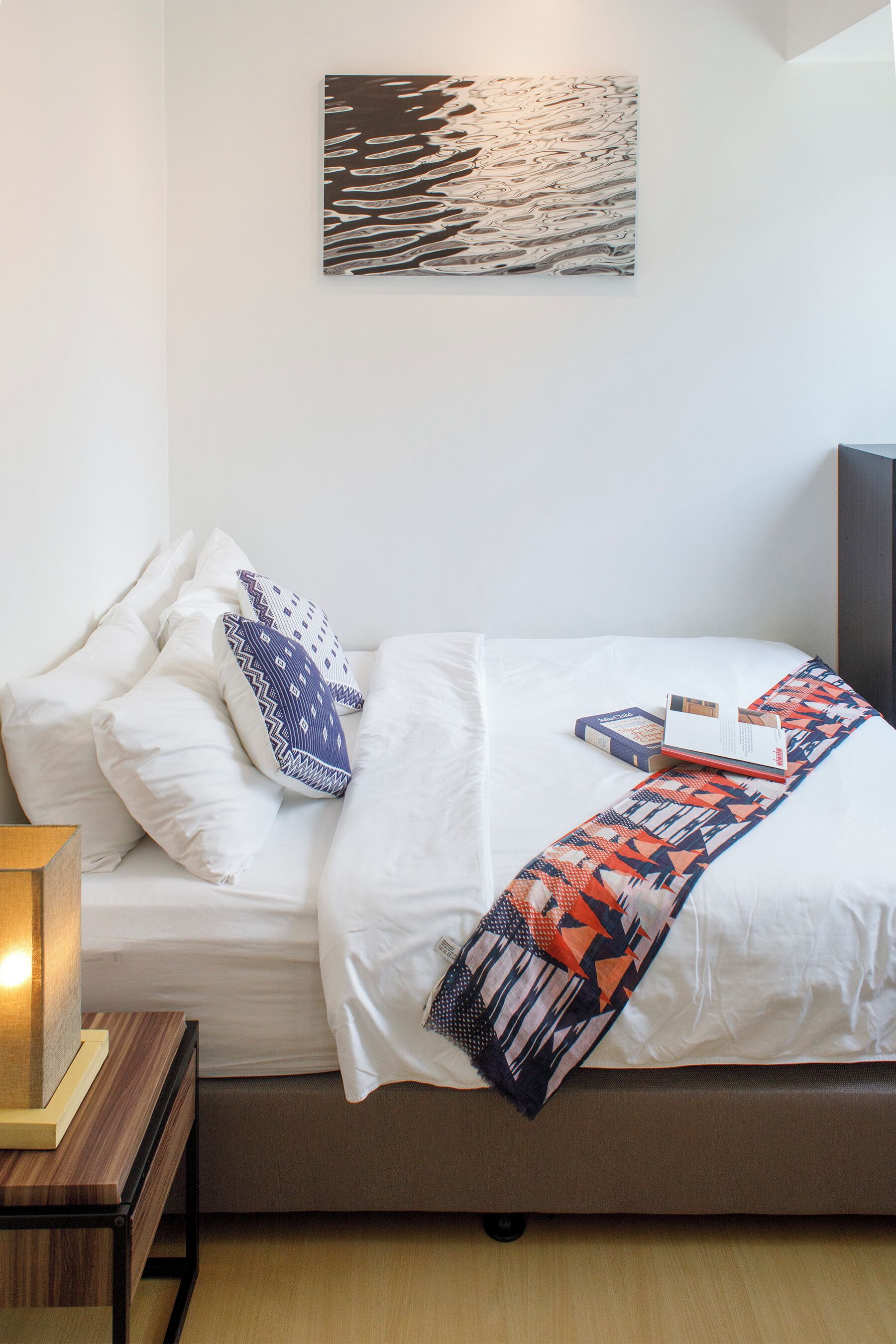 A neatly made bed with white bedding and a colorful throw blanket, a bedside table with a lamp, and a wall-mounted abstract painting.