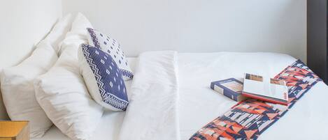 A neatly made bed with white bedding and a colorful throw blanket, a bedside table with a lamp, and a wall-mounted abstract painting.