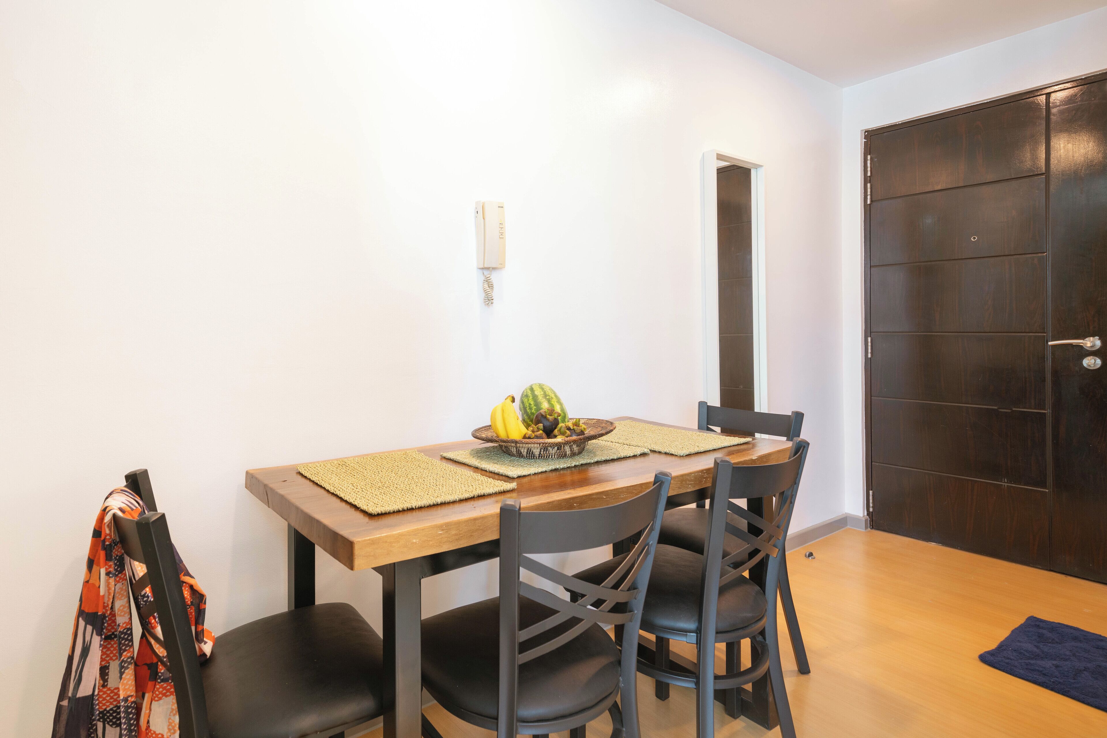 Un comedor con una mesa de madera, cuatro sillas negras, un cuenco de fruta y un teléfono en la pared.
