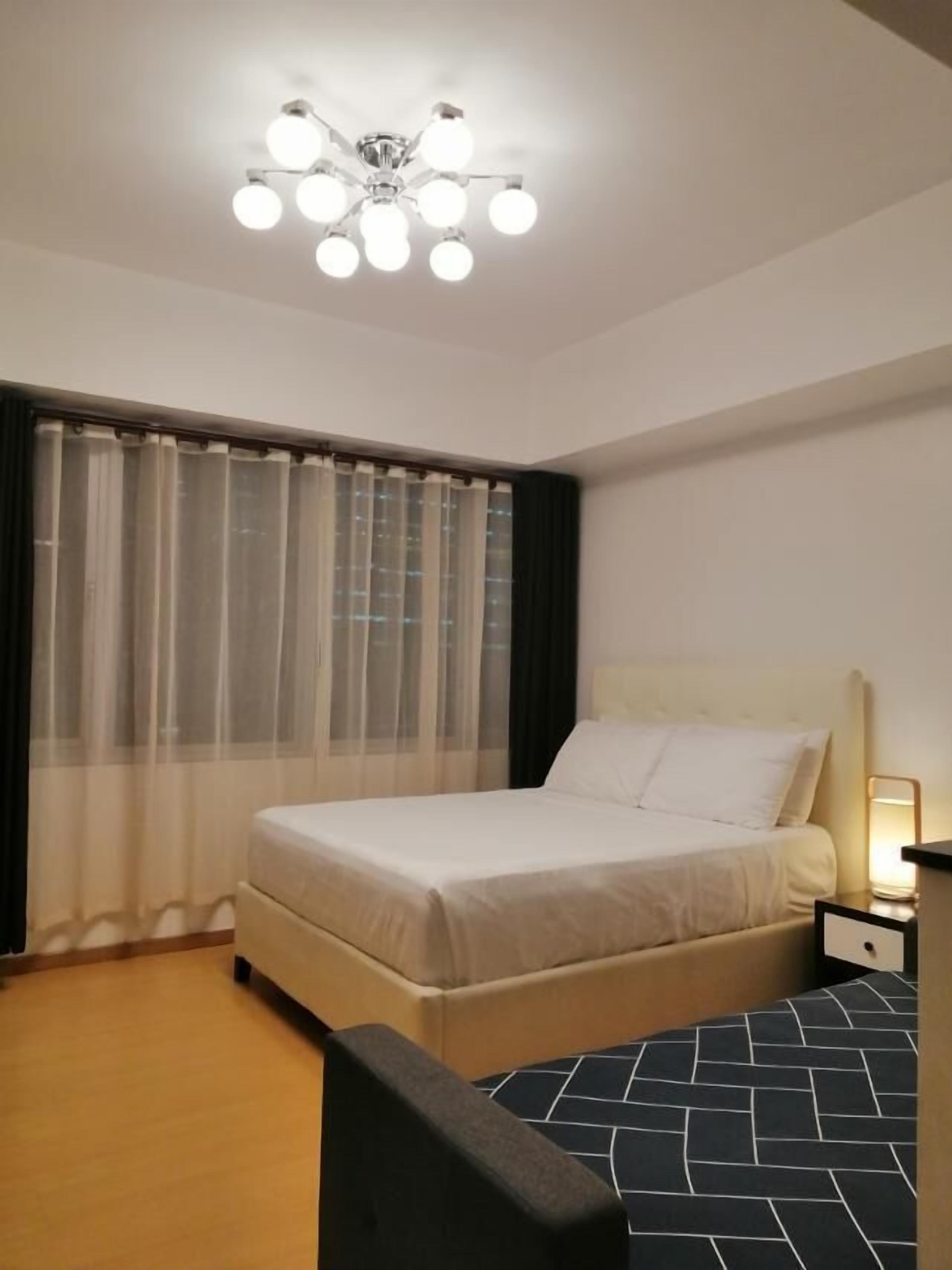 Ett hotellrum med en säng, ett sängbord, en stol, en taklampa och tunna, genomskinliga gardiner.