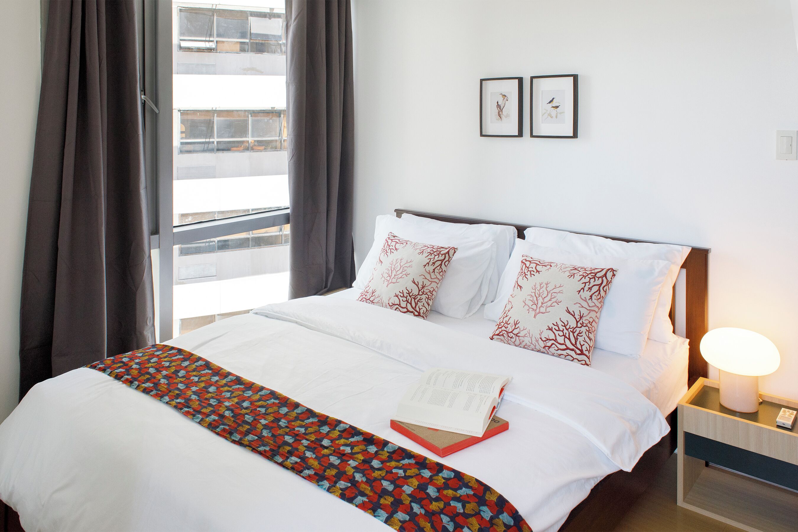 Una cama bien hecha con sábanas blancas y almohadas decorativas, una manta colorida y un libro sobre la cama. Hay una mesita de noche con una lámpara y un teléfono.