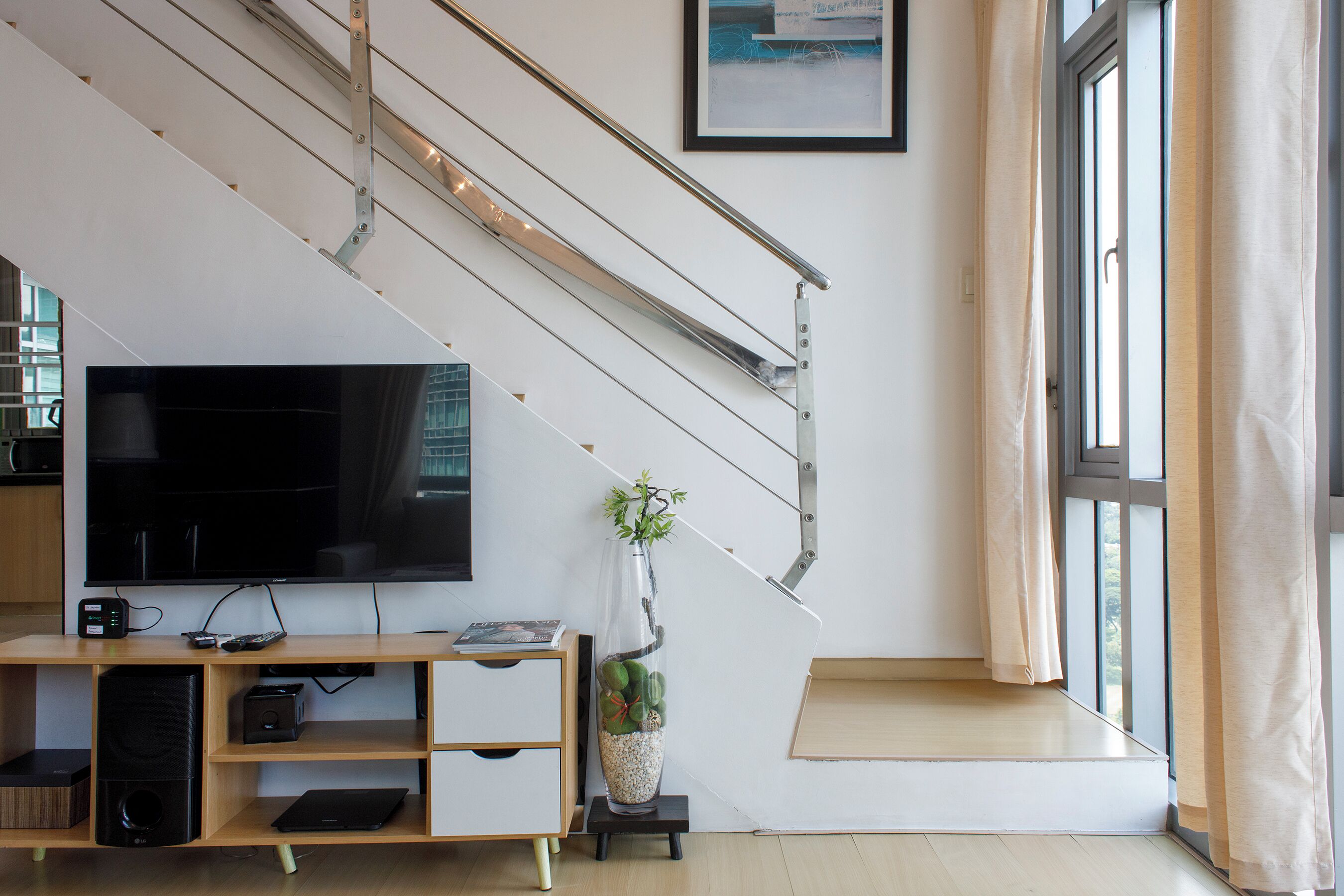 A modern living room with a flat-screen TV, wooden entertainment unit, and a staircase with a glass railing.