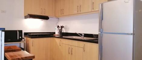 Een kleine keuken met houten kastjes, een witte koelkast, een magnetron en een klein houten tafeltje.