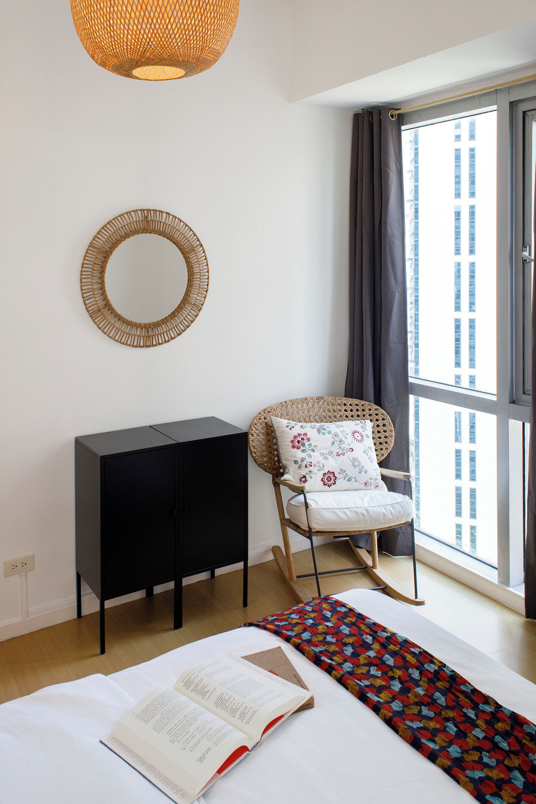 Un dormitorio con cama, un armario negro, un sillón de mimbre con cojín floral, un espejo redondo y un ventanal con cortinas.