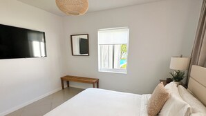 Ein Schlafzimmer mit einem großen Bett, einer Holzbank, einem Fenster mit Jalousien, einem am Wand montierten Flachbildfernseher und einem Nachttisch mit Lampe und Pflanze.
