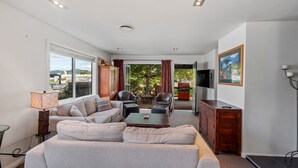 Una sala de estar moderna con sofá, mesa de centro y televisor. Se aprecia una vista al exterior a través de amplios ventanales.
