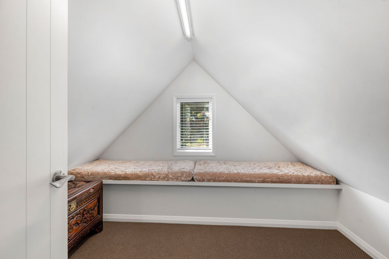 Ein kleines Schlafzimmer im Dachgeschoss mit schrägen Wänden, einem Fenster mit Jalousien, einer Bank und einer hölzernen Kommode.