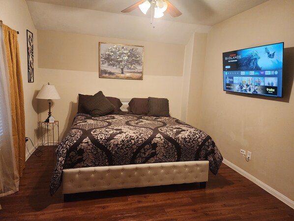 A bedroom with a large bed, a ceiling fan, a TV mounted on the wall, and a painting above the bed.