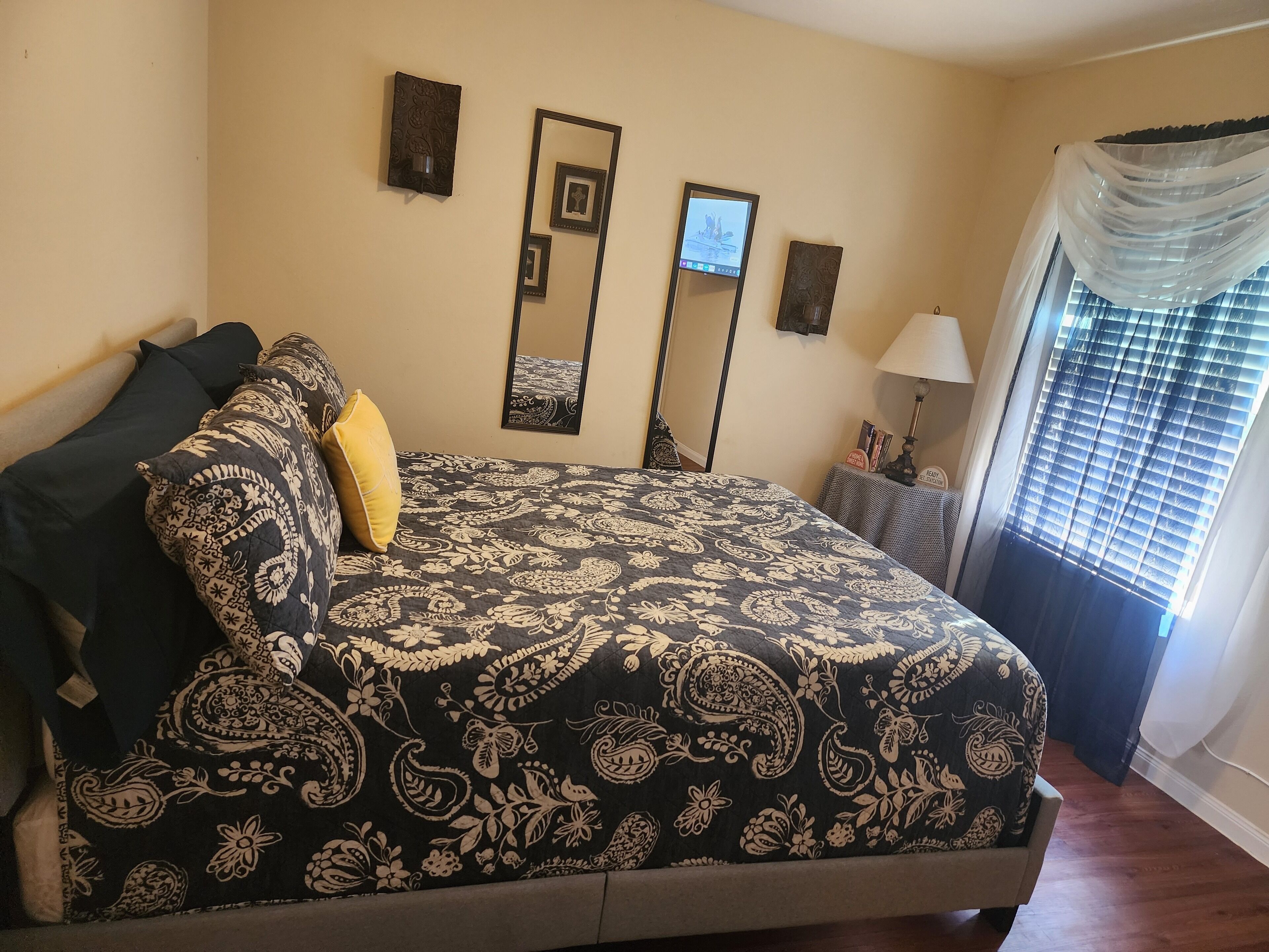 A bedroom with a bed, a nightstand, a mirror, and a window with curtains.