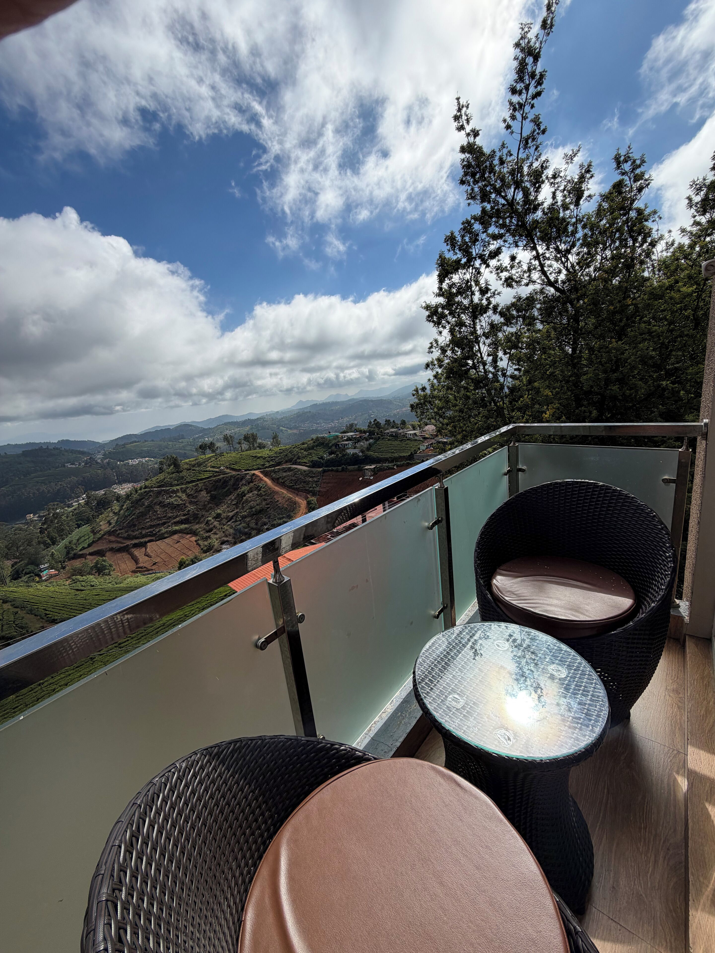A balcony with wicker chairs and a small table, overlooking a scenic landscape with hills and a river.