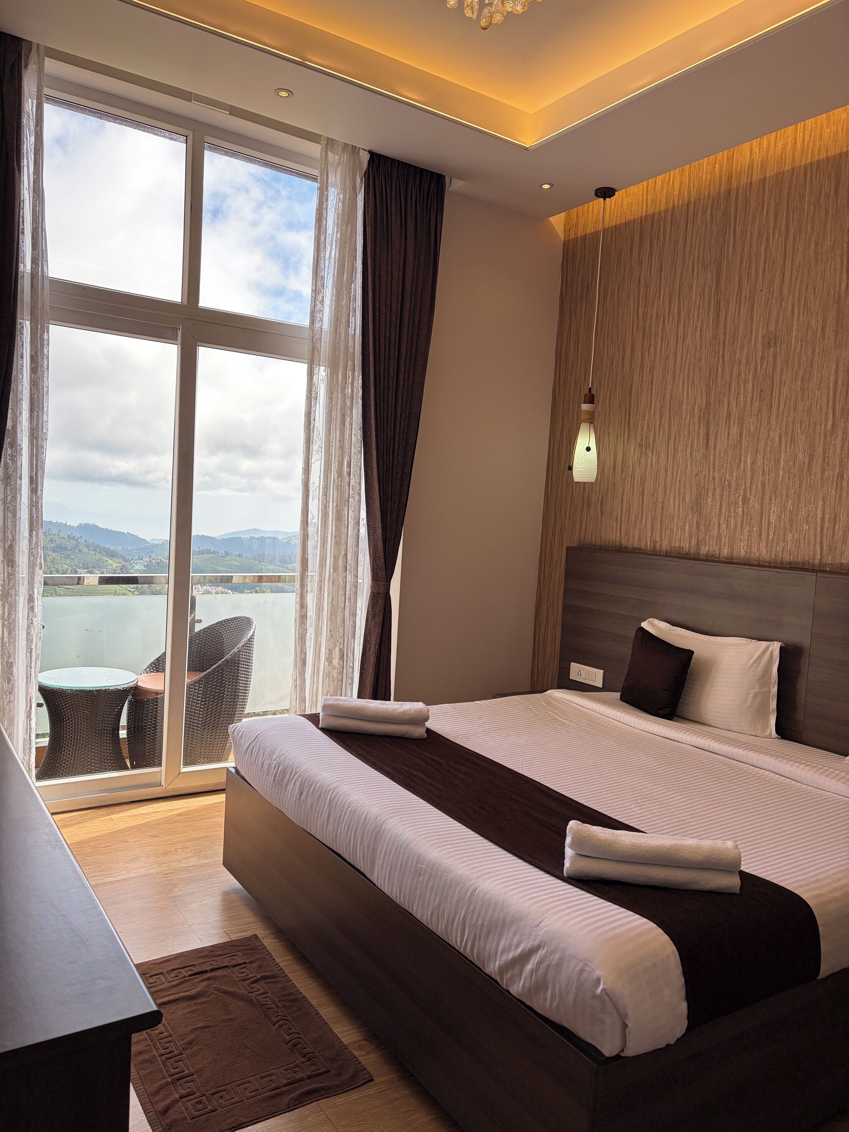 A modern hotel room with a large bed, a view of the outdoors, and a balcony with a table and chairs.