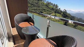 A balcony with wicker chairs and a table, overlooking a green landscape with hills and a clear sky.