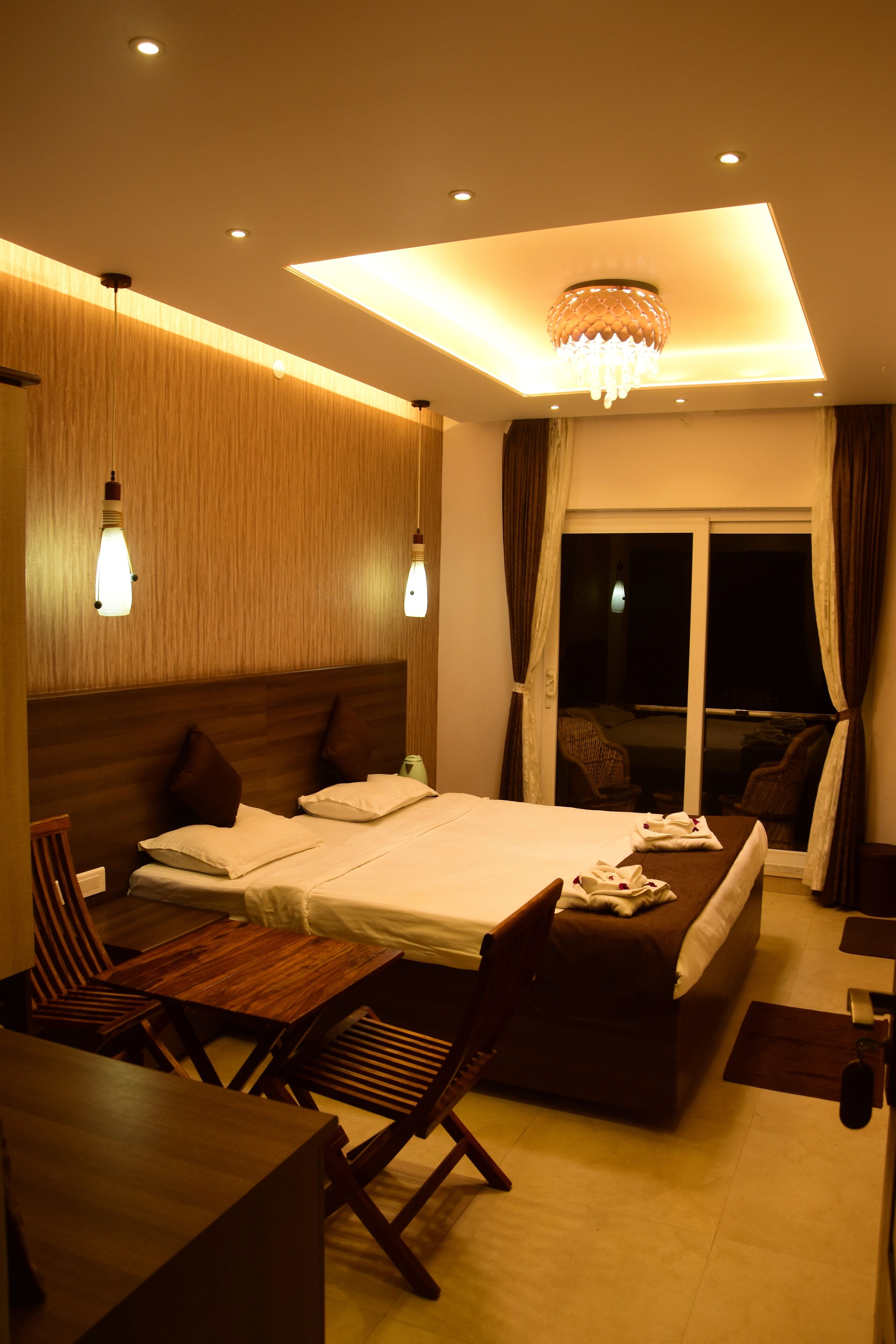 A modern hotel room with a large bed, a desk, and a chair. There is a window with curtains, and a chandelier on the ceiling.