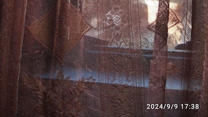 Ein Fenster mit einem gemusterten Vorhang, durch das Bäume und ein Gebäude sichtbar sind.