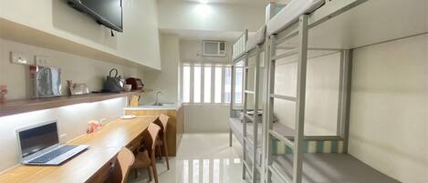 A dormitory room with bunk beds, a desk with a laptop, a TV mounted on the wall, and a window providing natural light.