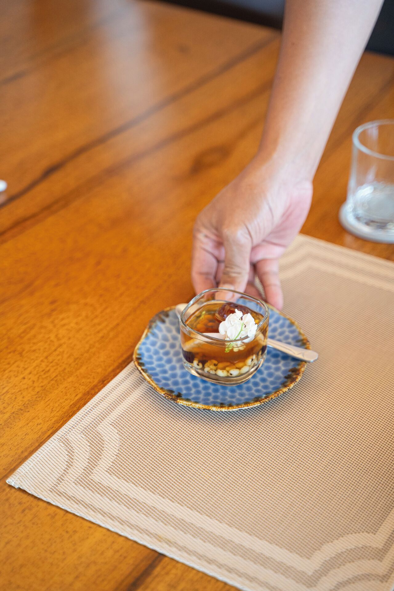 A hand holding a small glass cup with a spoon on a blue patterned coaster, placed on a beige placemat on a wooden table.