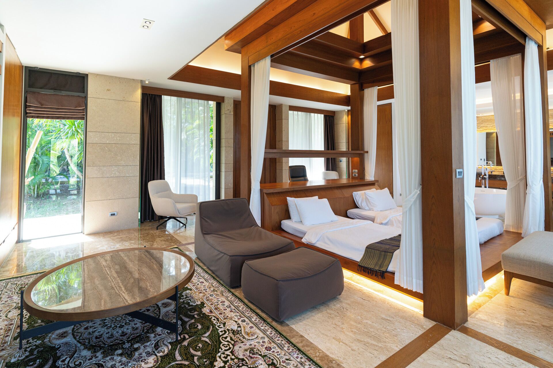 Una habitación de hotel moderna con una cama grande, un sofá, una mesita y una silla. Hay un ventanal con cortinas, vistas a zonas verdes y una estructura de techo de madera.