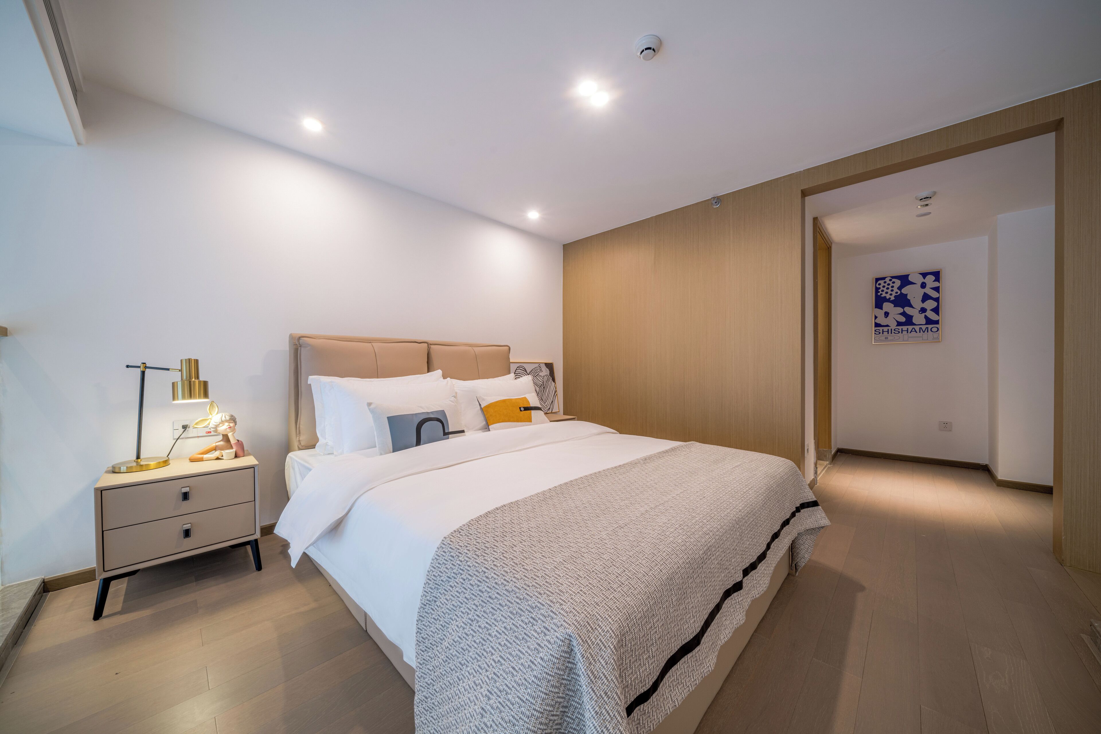 A modern bedroom with a large bed, a bedside table with a lamp, and a wall-mounted artwork.