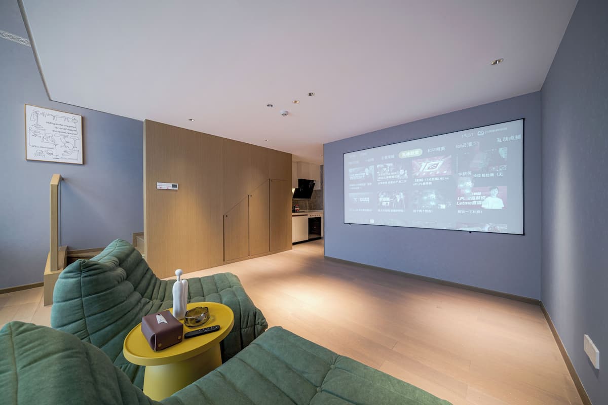 A modern living room with a large screen displaying a presentation, green upholstered seating, and a small yellow table with a remote control.