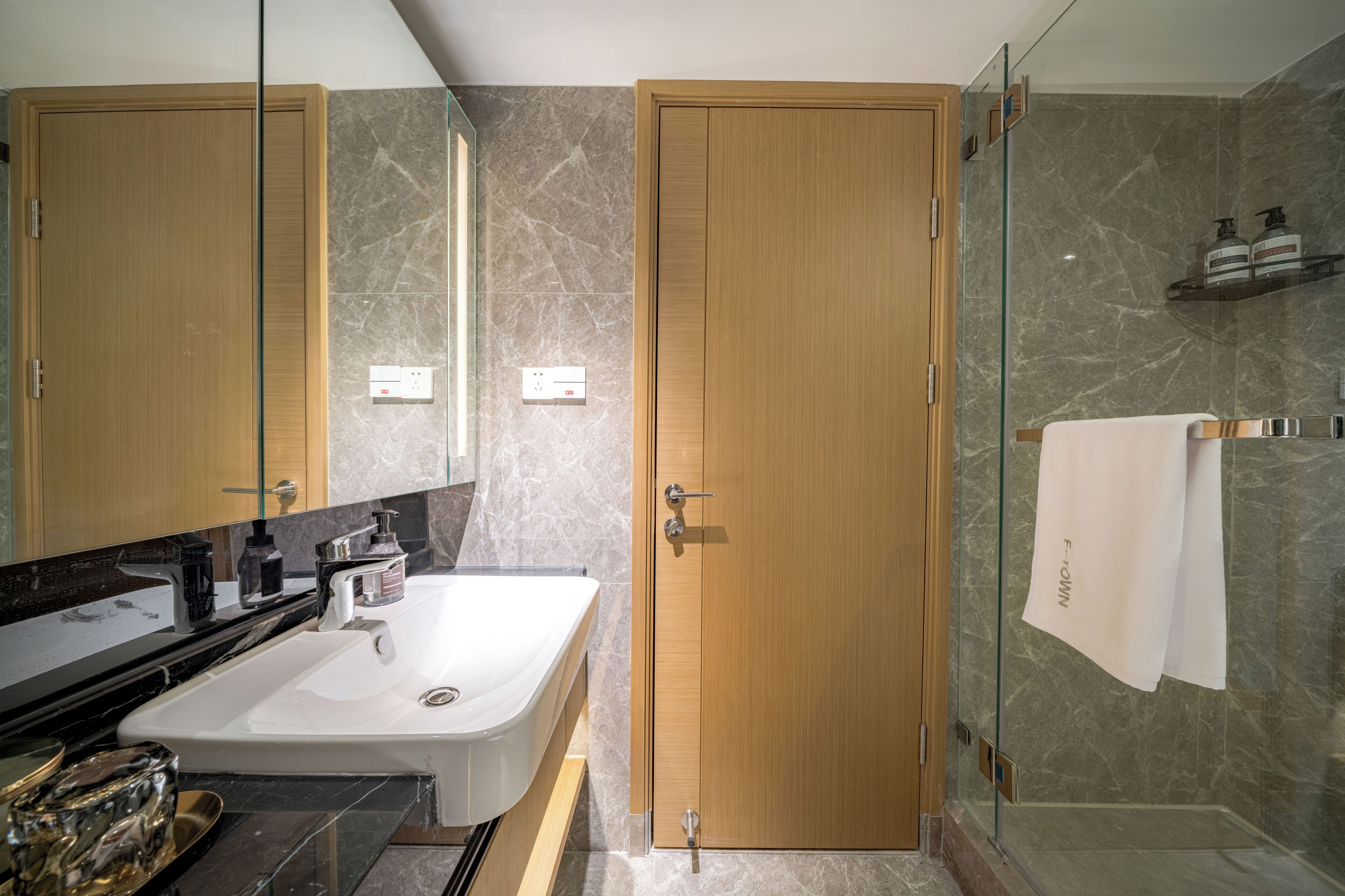 Un baño moderno con puerta de madera, cabina de ducha de vidrio y lavamanos blanco.